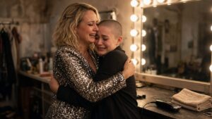 A young woman with a freshly shaved head smiling through tears as a blonde woman hugs her in a backstage dressing room
